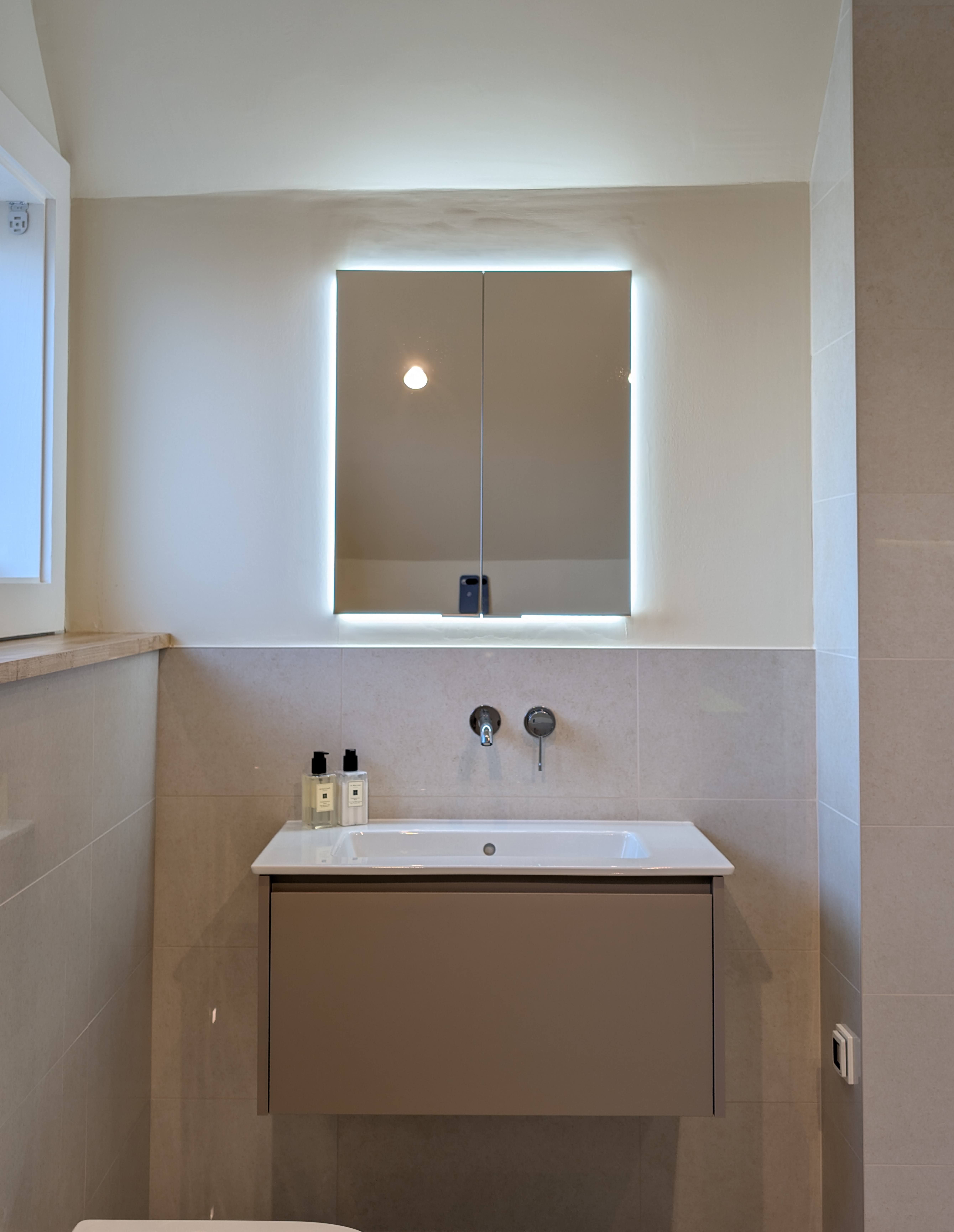 Wall-mounted basin below an illuminated mirrored cabinet